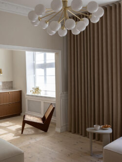 Luxaflex® stofgardiner i en mørk beige, der hænger i en stue, hvor der også er en lænestol.